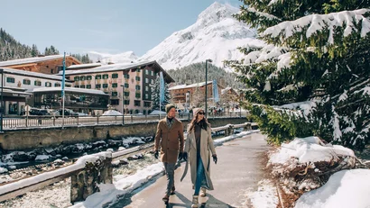 Paar spaziert im verschneiten Bergdorf mit Bergen und Tannen im Hintergrund