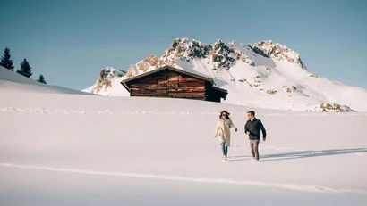 Paar läuft im Schnee vor einer Holzhütte mit schneebedeckten Bergen im Hintergrund