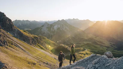 Zwei Wanderer steigen in den Bergen bei Sonnenuntergang einen steilen Hang hinauf