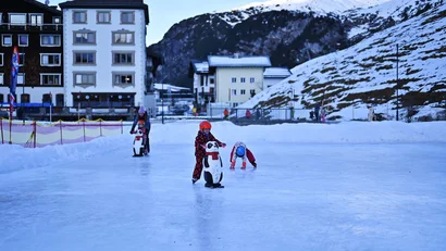 Kinder laufen mit Pinguin-Hilfen auf einer Eisbahn vor verschneiten Bergen und Häusern