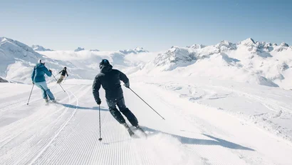 Drei Skifahrer auf präparierter Piste vor schneebedeckten Bergen